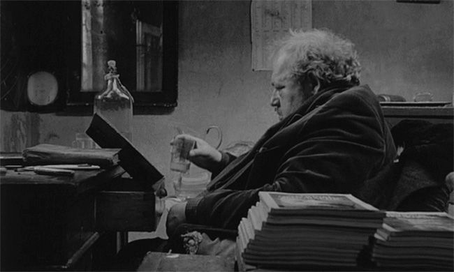 Mientras observa por la ventana y escribe, el doctor bebe alcohol con frecuencia. Precisamente lo único que le puede hacer salir de casa, en dirección a la taberna del pueblo, es quedarse sin alcohol. ¿”Ayuda” para “aligerar” y “aclarar las ideas” en la dificultad del proceso creativo, simplemente “enfermedad” o ambas cosas a la vez? | Análisis cinematográfico en Cultugrafía. Difusión y crítica cultural. Cine y películas de Béla Tarr.