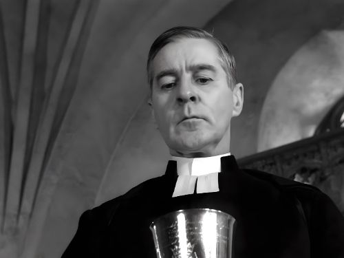 La imagen muestra a un hombre vestido con sotana, mirando hacia abajo con una expresión seria. Está sosteniendo un cáliz de plata en su mano. La imagen está en blanco y negro y tiene una atmósfera solemne y religiosa.